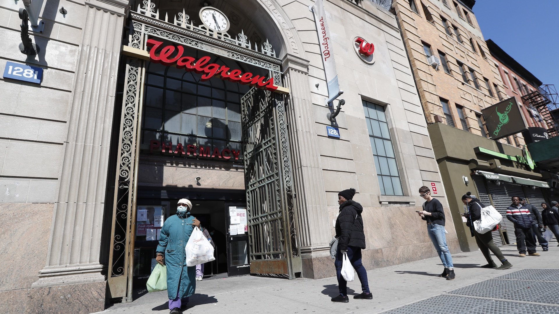 In this March 26, 2020 file photo, a woman leaves a Walgreens pharmacy that is enforcing social distancing rules by allowing only a certain numbers of people in as customers line up on the sidewalk outside the store in New York. The coronavirus pandemic pushed the drugstore chain to a .7-billion loss in its fiscal third quarter, as customers stayed home to avoid the virus or consolidated their shopping to grocery stores. (AP Photo/Kathy Willens, File)