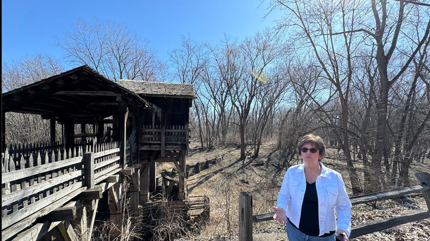 New Salem Lincoln League President Gina Gillmore-Wolter stands in front of the Grist Mill at the New Salem Historic Site on March 10, 2025. The mill’s drive shaft has been lying in the weeds near the structure since 2016 after it was dislodged due to flooding. (Athan Yanos / Medill Illinois News Bureau)