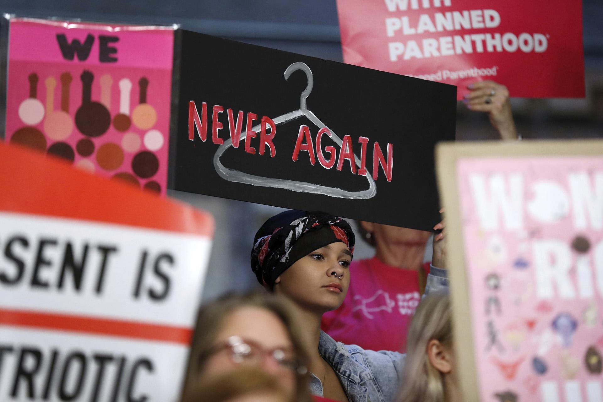 In this Tuesday, May 21, 2019 file photo, August Mulvihill, of Norwalk, Iowa, center, holds a sign depicting a wire clothes hanger during a rally at the Statehouse in Des Moines, Iowa, to protest recent abortion bans. (AP Photo / Charlie Neibergall)