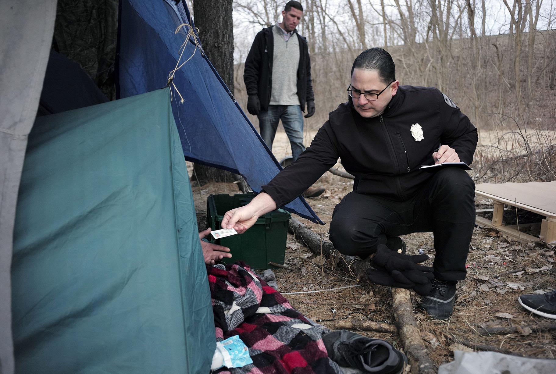 In this Tuesday, Feb. 12, 2019 file photo, Worcester Police Officer Angel Rivera, right, returns a license to an unidentified man as Rivera asks if he has been tested for Hepatitis A at the entrance to a tent where the man spent the night in a wooded area, in Worcester, Mass. (AP Photo / Steven Senne)
