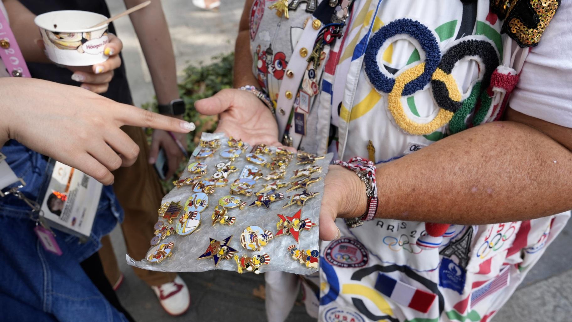 Vivianne Robinson lets a passerby choose one of the pins Robinson collected from USA Olympics 1984, during 2024 Summer Olympics, in Paris, France, Tuesday, July 30, 2024. The Olympics superfan has attended seven Games over the span of 40 years. (Lujain Jo / AP Photo)