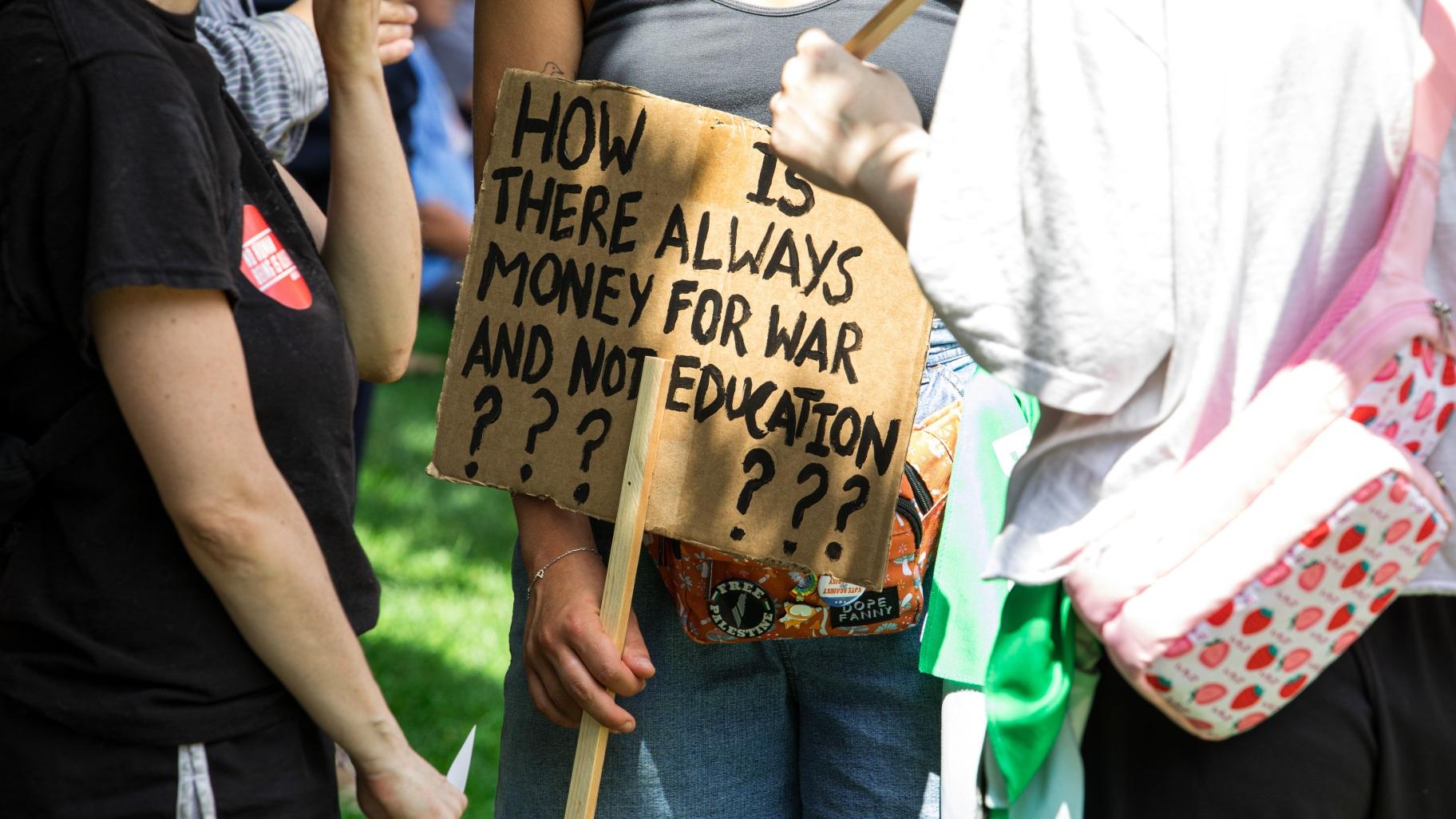 A protester holds a sign that reads “How is there always money for war and not education??????” moments before the march in downtown Milwaukee begins on Monday, July 15, 2024. (Michael Izquierdo / WTTW News)