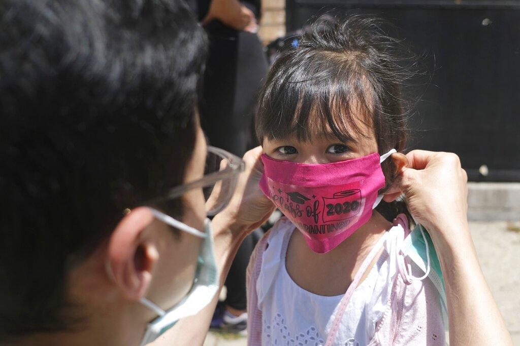 In this June 10, 2020, file photo, Olivia Chan’s father helps her with a new mask she received during a graduation ceremony for her Pre-K class in front of Bradford School in Jersey City, N.J. (AP Photo / Seth Wenig, File)