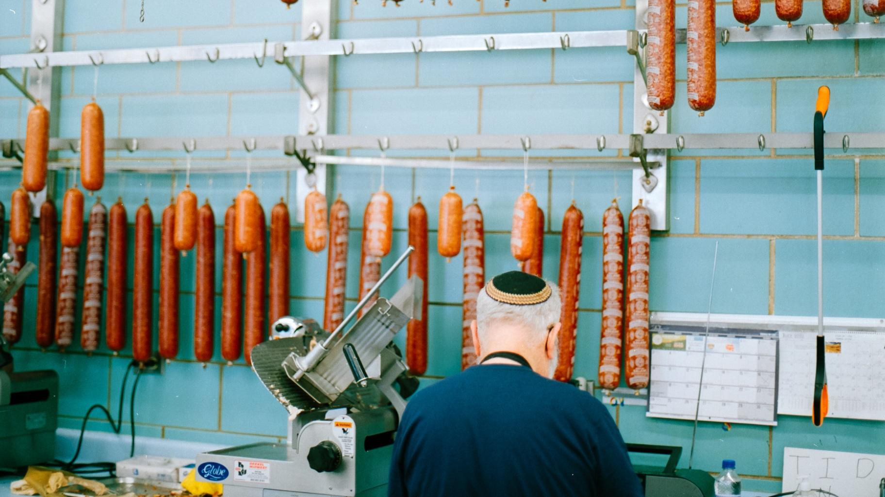 A butcher prepares an order as beef salamis dangle above him at Romanian Kosher Sausage Co. in Chicago, 2022. (Max Abrams)