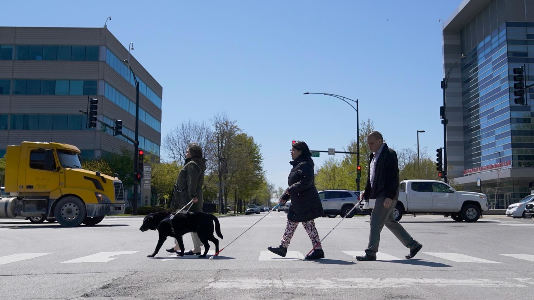 FILE - Maureen Reid, left, and her guide dog, Gaston, cross the intersection of Wood Street and Roosevelt Avenue with Sandy Murillo, center, and Geovanni Bahena, relying on an audible signal for the blind, on April 26, 2023, in Chicago. (Charles Rex Arbogast / AP Photo, file)
