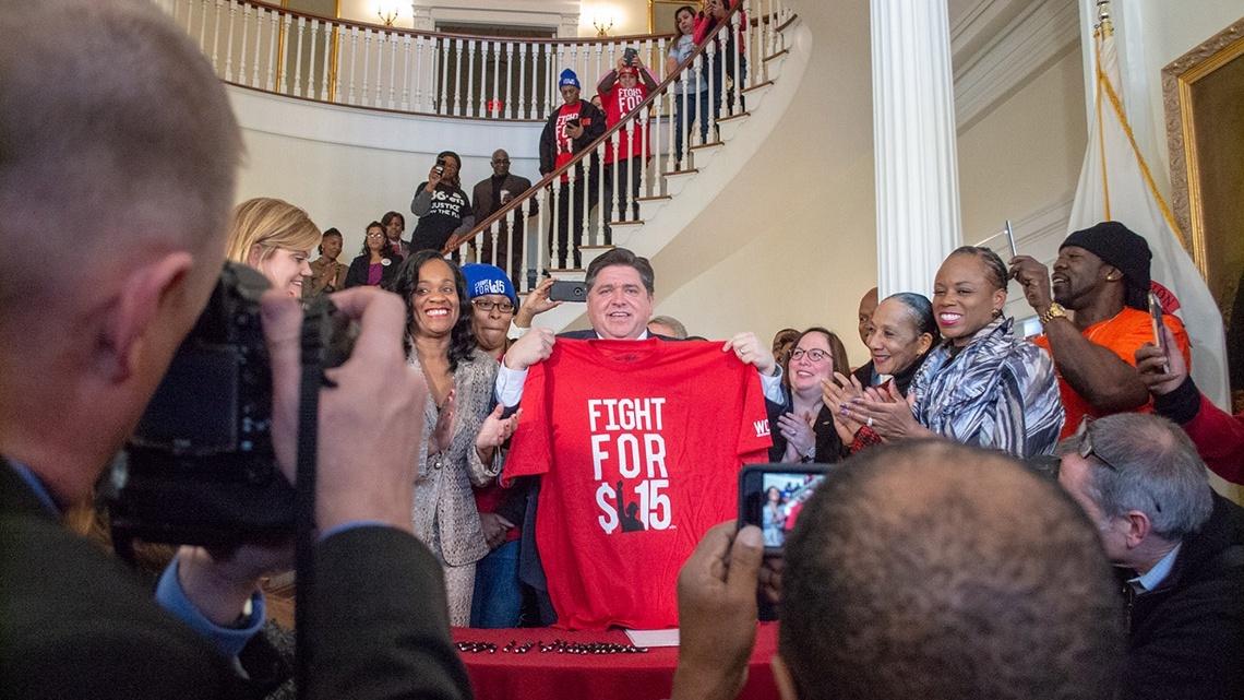 Gov. JB Pritzker holds a shirt commemorating the movement to raise the minimum wage to 5 after signing the bill on February 20, 2019. The wage hits 5 in 2025. (Capitol News Illinois photo by Jerry Nowicki)