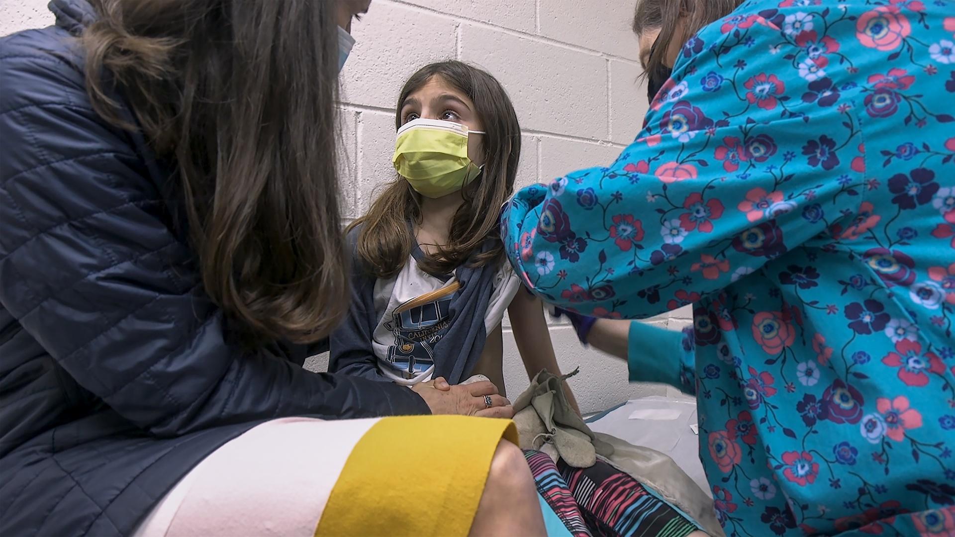 In this Wednesday, March 24, 2021 image from video provided by Duke Health, Alejandra Gerardo, 9, looks up to her mom, Dr. Susanna Naggie, as she gets the first of two Pfizer COVID-19 vaccinations during a clinical trial for children at Duke Health in Durham, N.C. (Shawn Rocco/Duke Health via AP)
