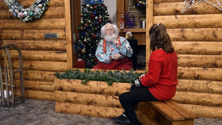 Santa, Sid Fletcher, sits behind a glass barrier as he hears Kendra Alexander of St. James, Minn., during her visit Monday, Nov. 15, 2021, at The Santa Experience at the Mall of America in Bloomington, Minn. (AP Photo/Jim Mone)