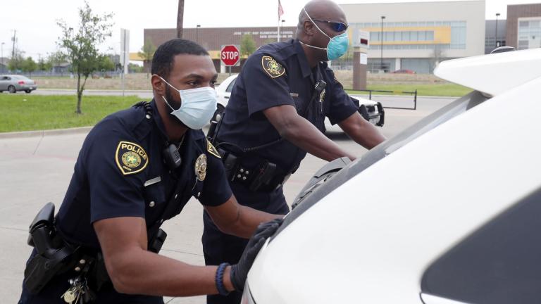 In this April 9, 2020, photo, amid concerns of the spread of COVID-19, Dallas ISD police officers Mylon Taylor, left, and Gary Pierre push a car that ran out of gas while waiting in line for the weekly school meal pick up for students in Dallas. The coronavirus pandemic that has crippled big-box retailers and mom and pop shops worldwide may be making a dent in illicit business, too. (AP Photo/LM Otero)