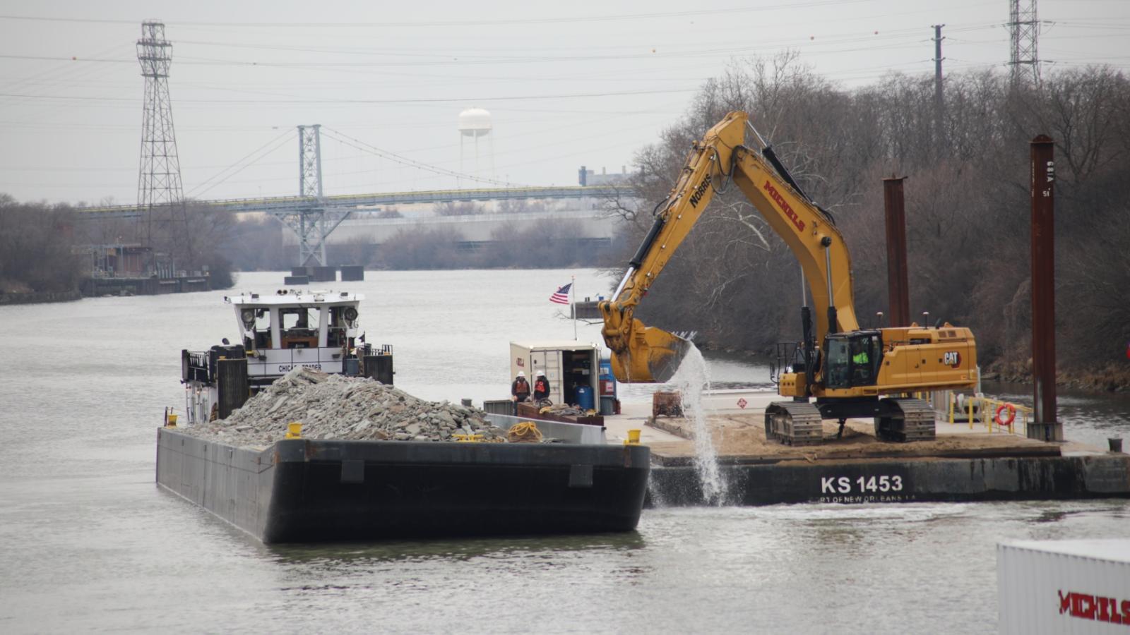 A contractor working on the Brandon Road Interbasin Project uses an excavator on a barge to gather broken bedrock from the bottom of the Illinois Waterway and place it on another barge for removal from the site. (U.S. Army Corps of Engineers)